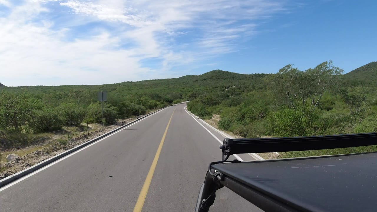 Opening view of a Los Cabos two-lane road running straight toward low green hills, with the vehicle edge visible at lower right and no visible people.
