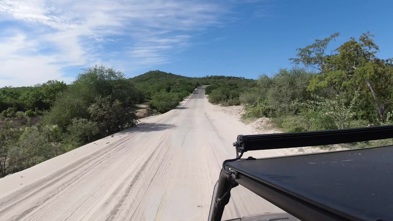 Forward view from a vehicle on a paved road in Los Cabos, Mexico, with sandy shoulders, green scrub, distant hills, blue sky, and no visible people.