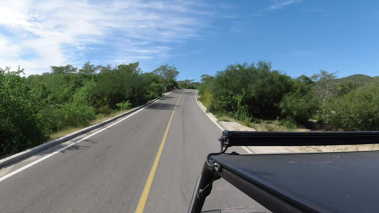 Paved road ahead with green brush and vehicle roof in foreground.