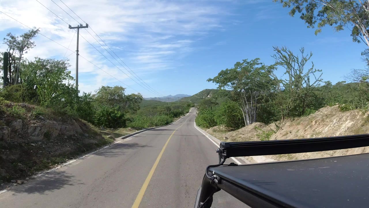 Forward view from a black vehicle on a two-lane road in Los Cabos, Mexico, with green brush, utility lines, distant hills, blue sky, and no visible people.