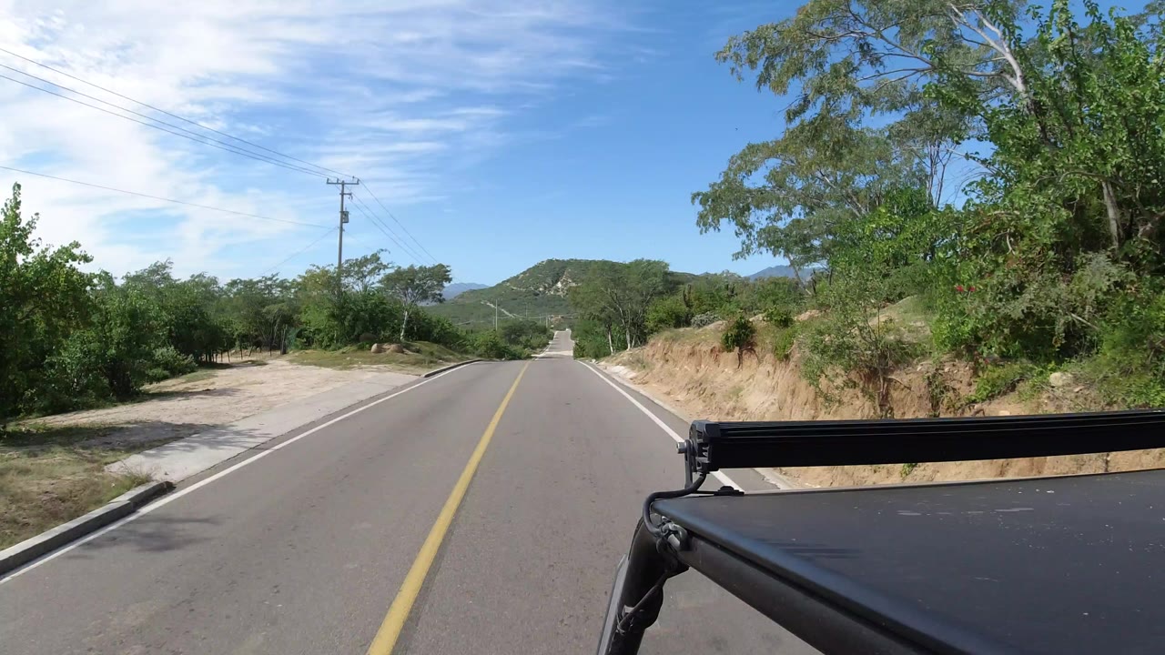 Forward vehicle POV on a two-lane road in Los Cabos, Mexico, with a yellow center line, green brush, sandy shoulder, distant hills, blue sky, and no visible people.
