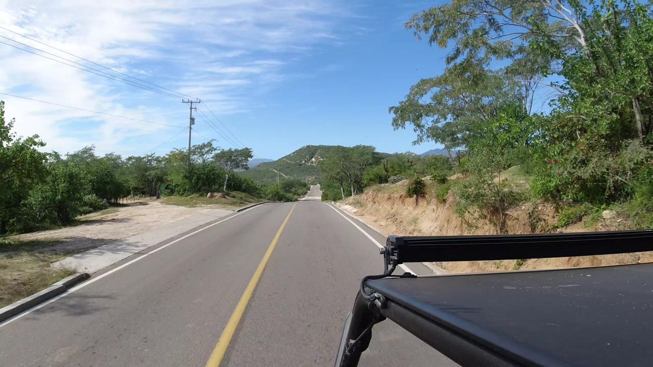 Vehicle-mounted view down a straight Los Cabos road, with trees close to the right shoulder, utility lines overhead, low hills ahead, and no visible people.