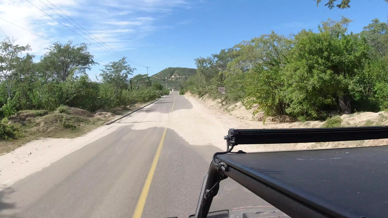 Forward vehicle view on a Los Cabos, Mexico road with a yellow center line, sandy roadside edges, dense green trees, low hills ahead, and no visible people.