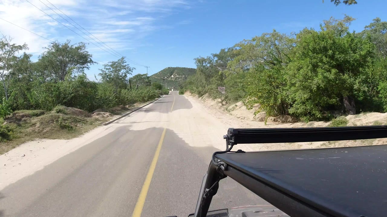 A centered Los Cabos road continues toward low hills, with green vegetation on both sides, pale sandy shoulders, visible utility lines, and no visible people.