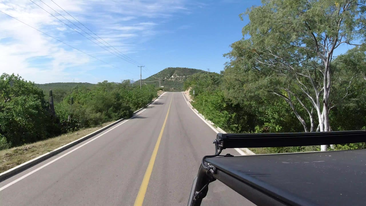Opening view from a vehicle on a Los Cabos, Mexico road, with a yellow center line, green roadside vegetation, distant hills, and no visible people.