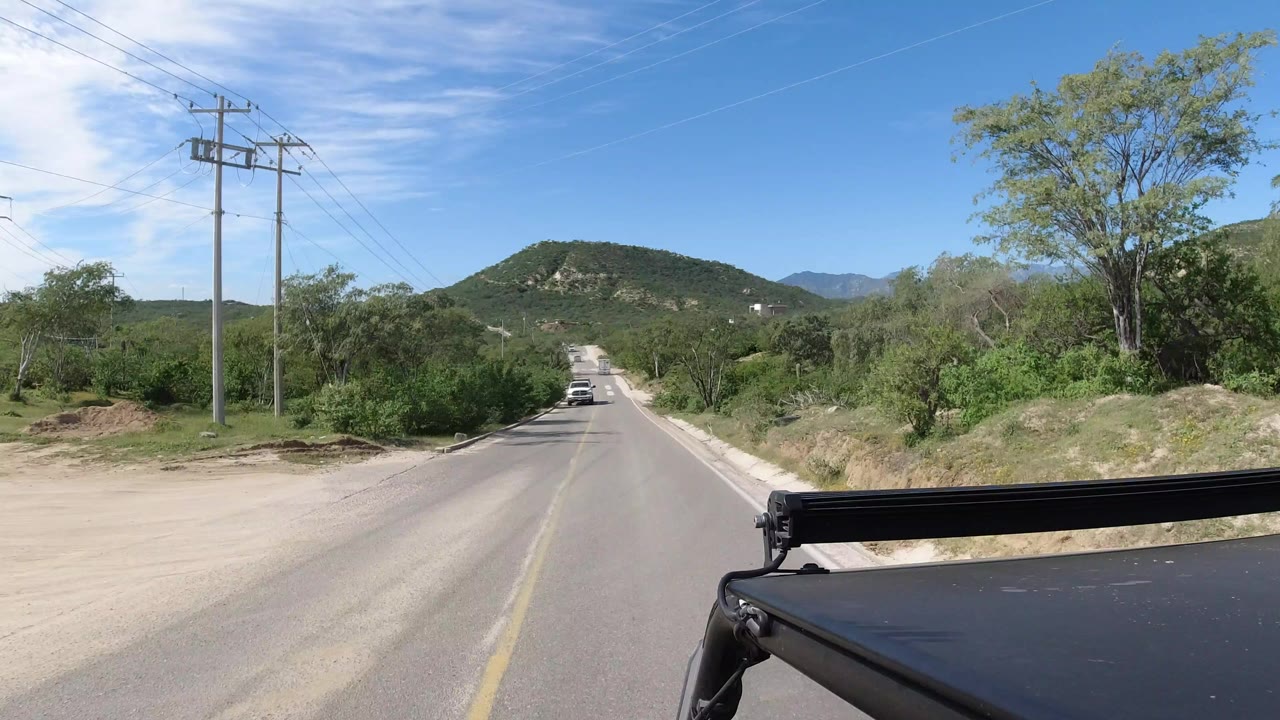 Forward view down a two-lane road with hills, power poles, and vehicle hardware in the foreground.