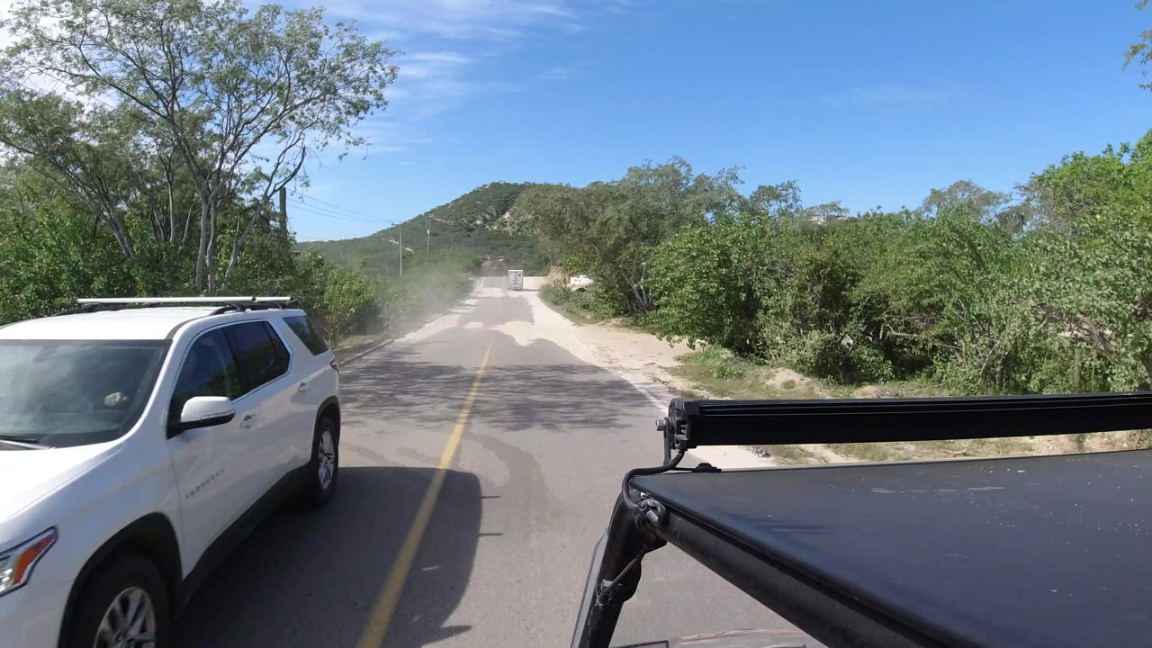 The white SUV remains near the left edge as the Los Cabos road runs ahead between green desert brush, sandy shoulders, low hills, and the filming vehicle roof, with no visible people.