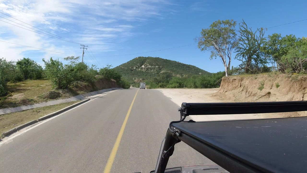 Forward road view in Los Cabos, Mexico, with a yellow center line, low green hills ahead, roadside scrub, and black vehicle hardware in the foreground, with no visible people.