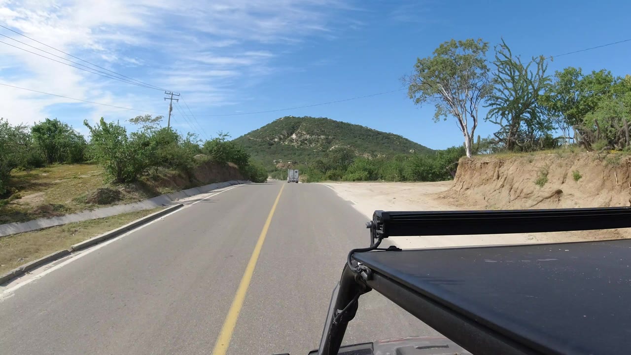 A forward-facing Los Cabos road view shows sandy roadside banks, green brush, utility lines, distant hills, and black vehicle hardware along the lower right, with no visible people.