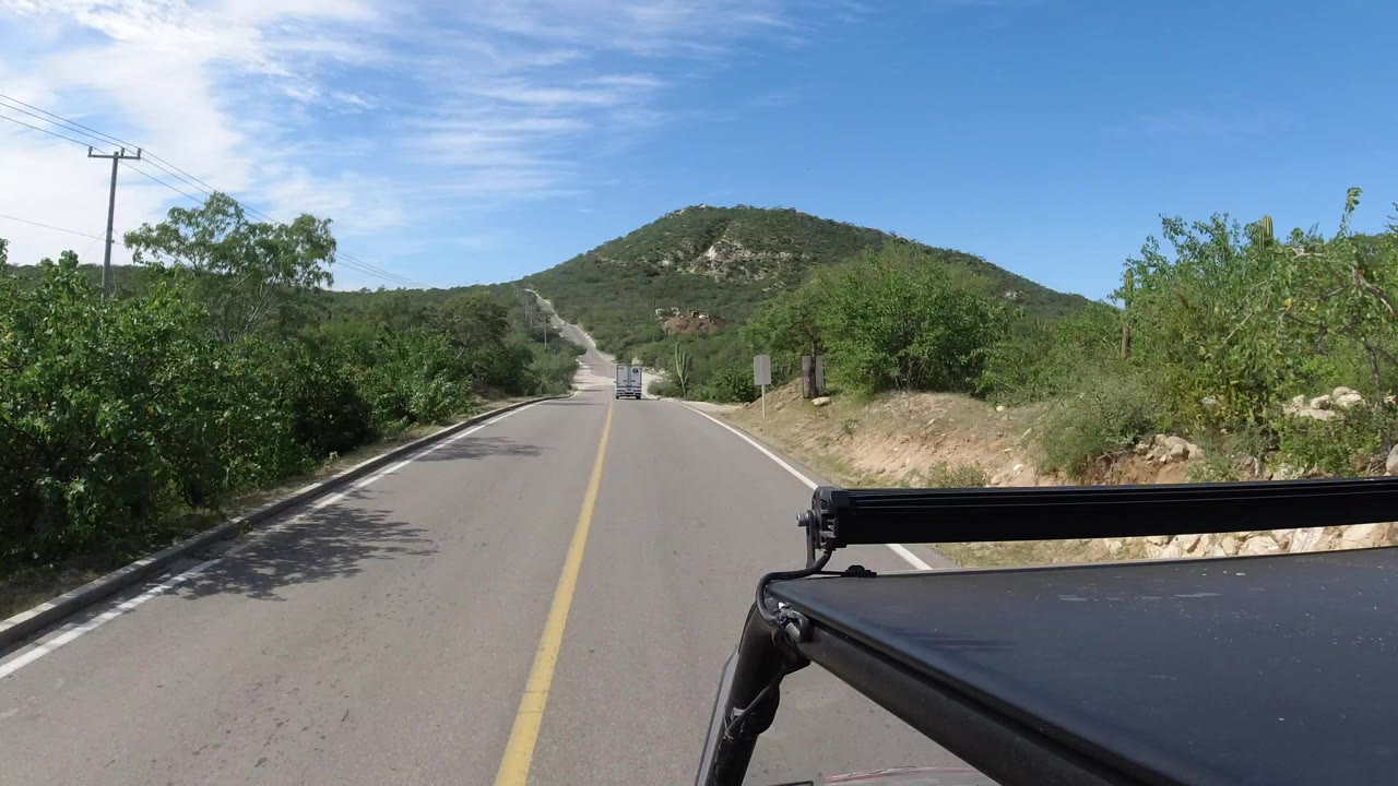 Forward vehicle view on a narrow Los Cabos road with shrubs, utility poles, a green hill ahead, blue sky, and no visible people.