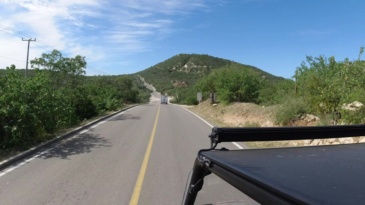 A slightly farther view along the Los Cabos road shows the centerline, roadside brush, utility wires, distant vehicle, and no visible people.