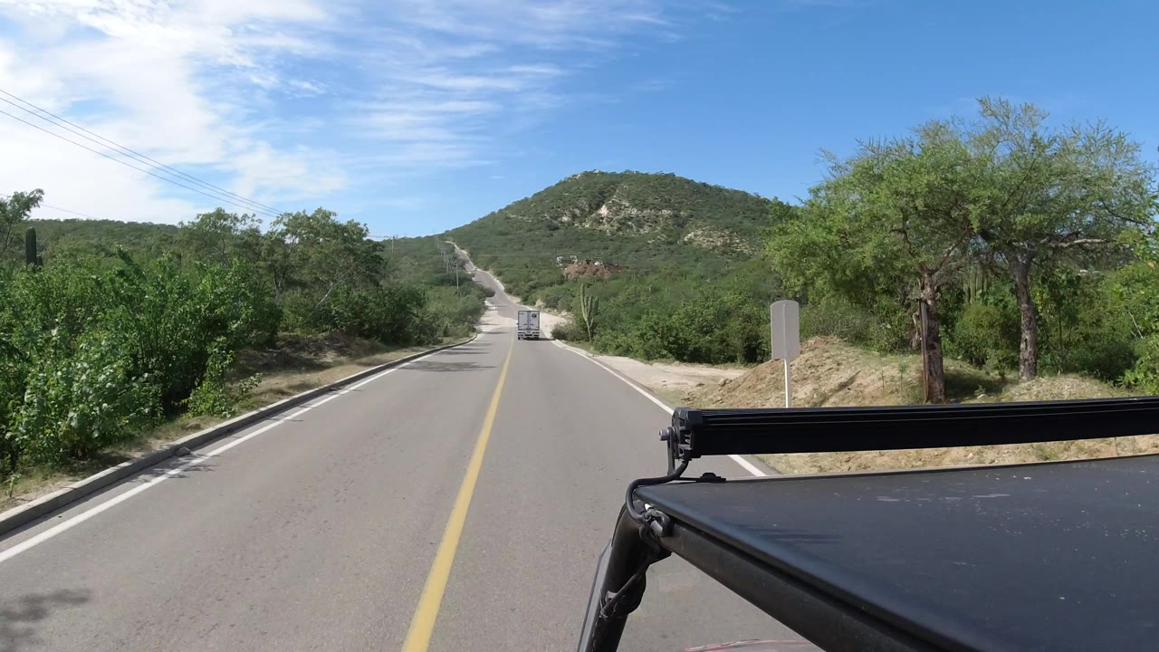 Road curves gently through green scrub with a hill in the distance.