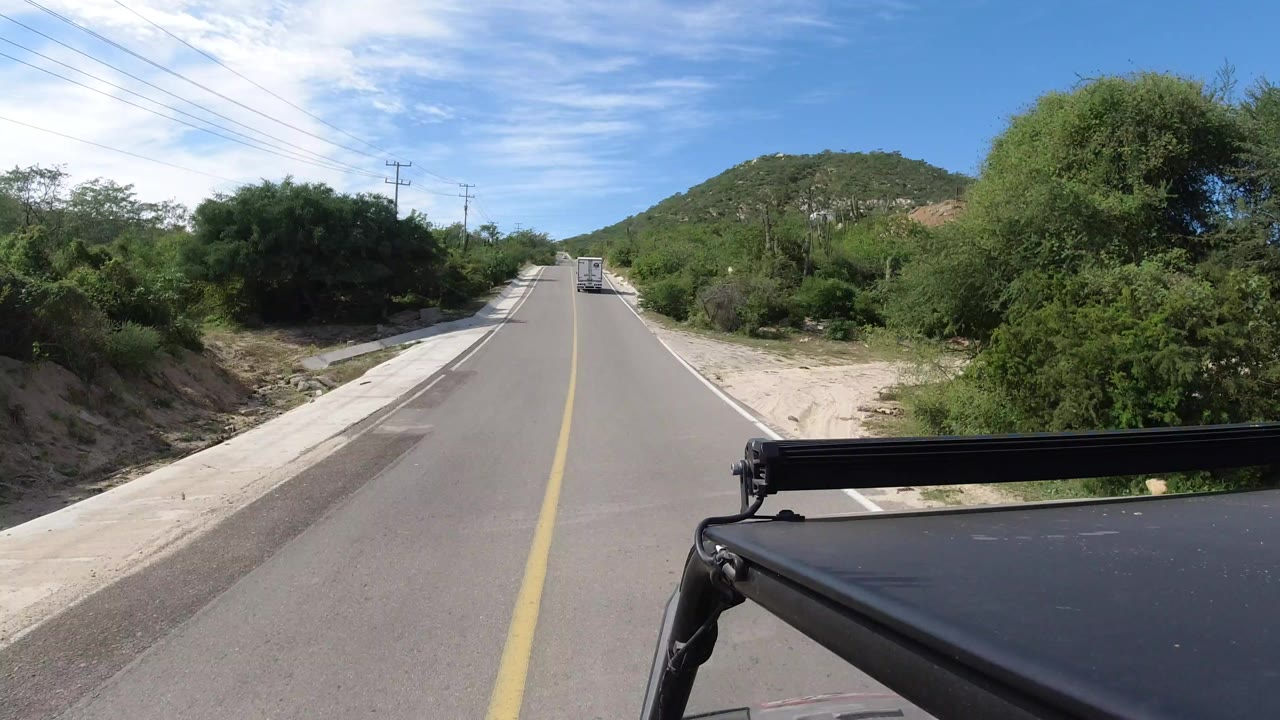 Opening view from a moving vehicle on a straight Los Cabos road, with a yellow centerline, roadside scrub, utility poles, a low green hill ahead, and no visible people.