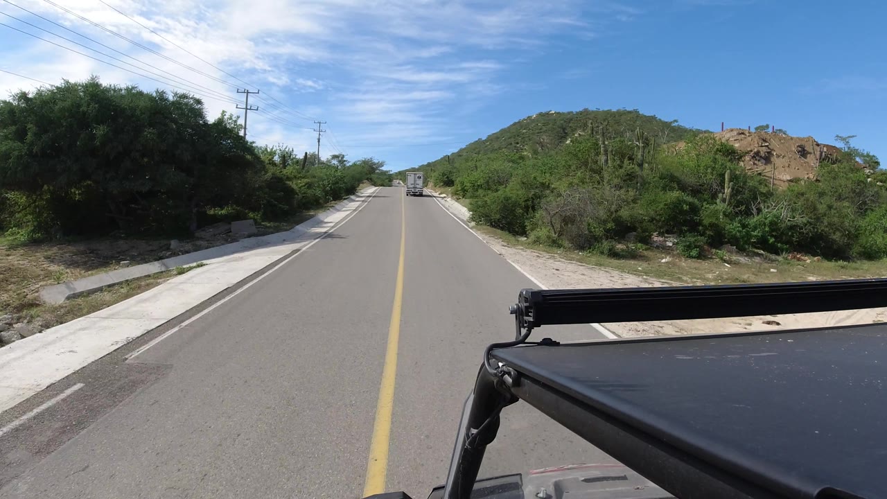 Road curving gently past shrubs and a hillside, with vehicle roof visible at lower right.
