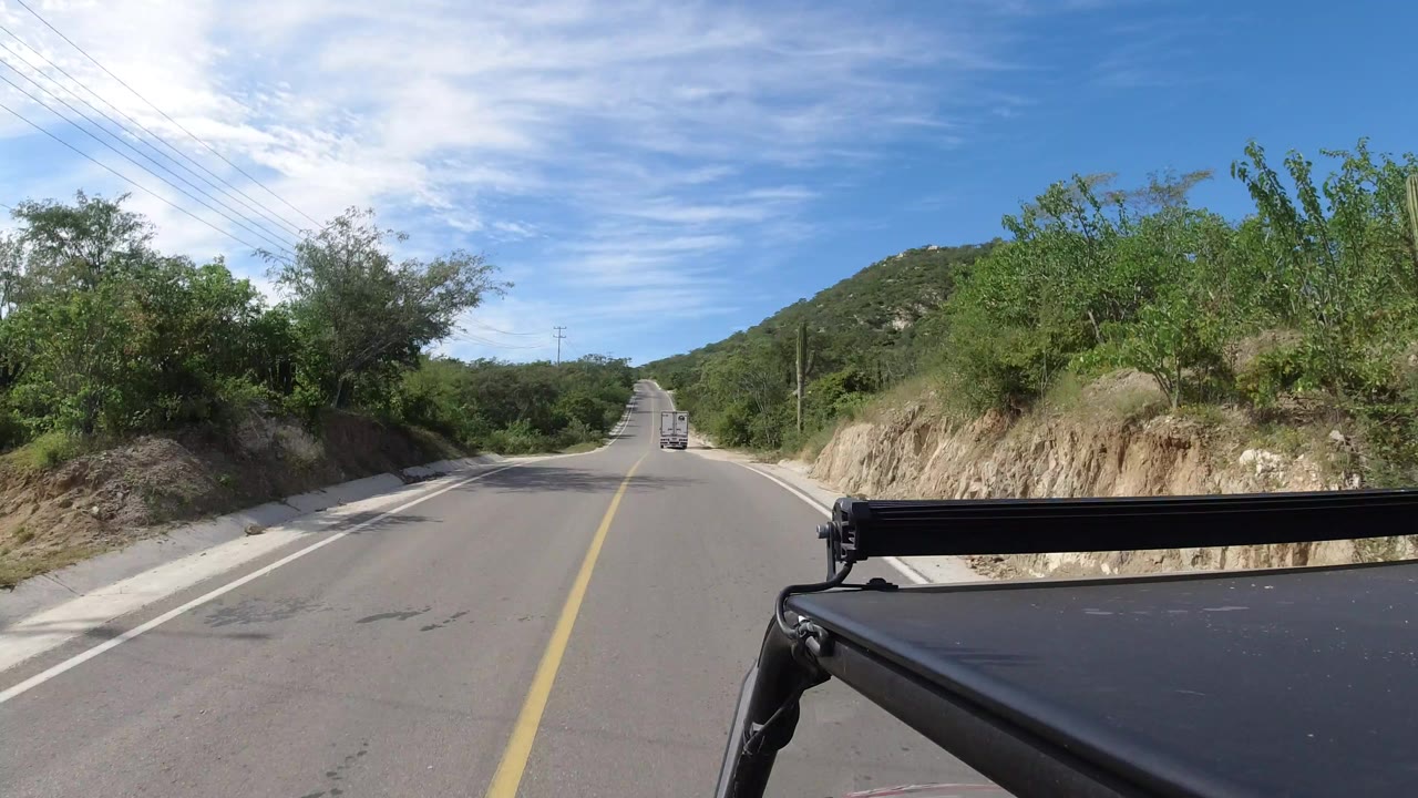 Forward vehicle POV on a paved road in Los Cabos, Mexico, with a distant vehicle ahead, green brush, low hills, utility lines, blue sky, and no visible people.