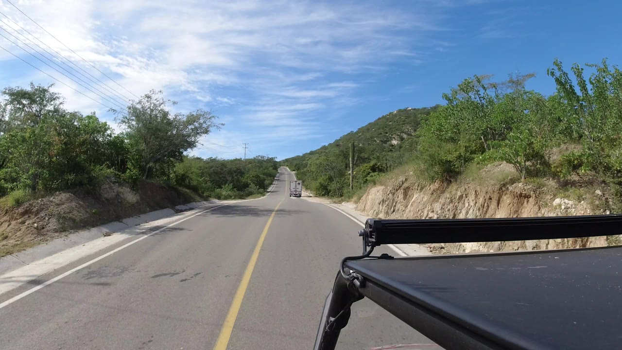 Los Cabos, Mexico road POV with the centerline leading into low hills, dense green roadside vegetation, a vehicle far ahead, and no visible people.