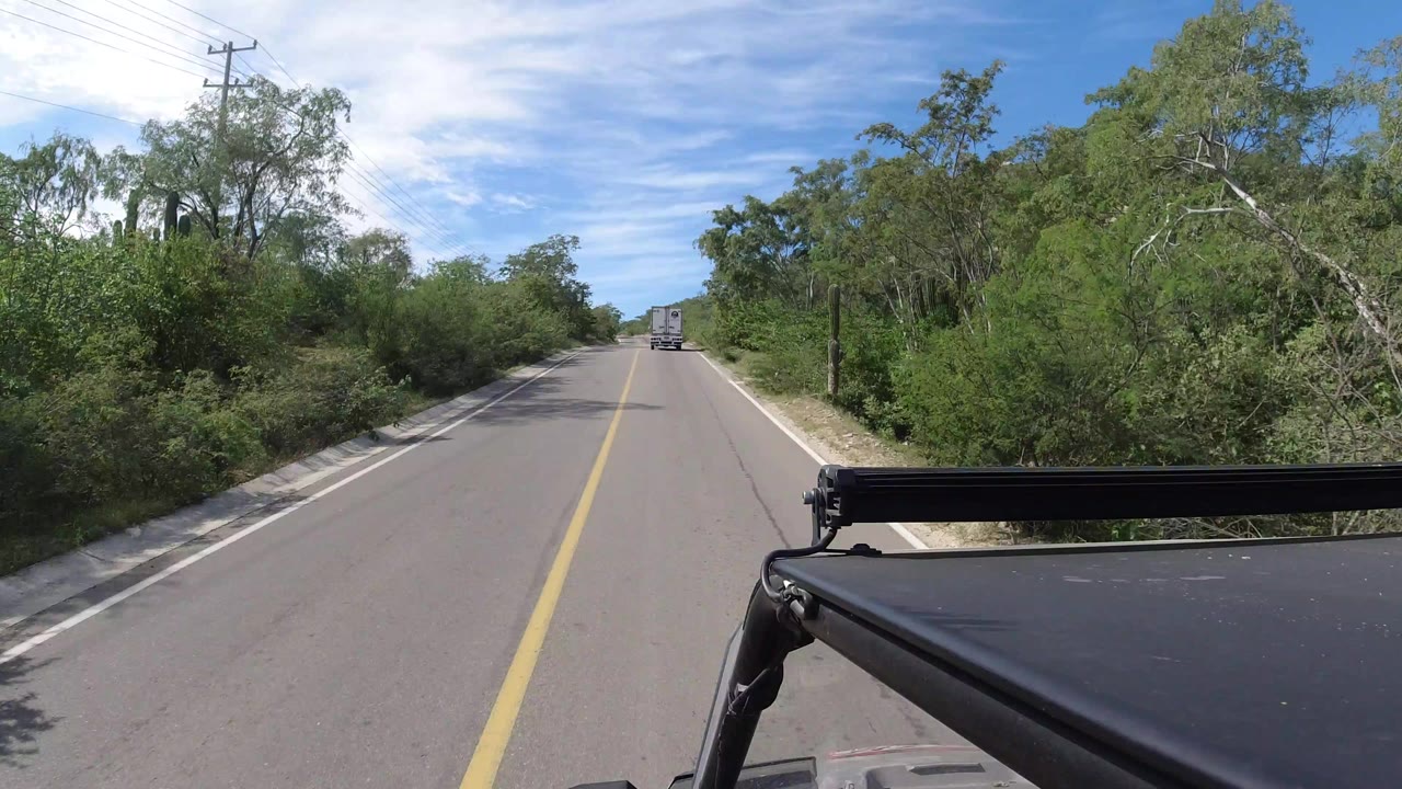 Forward-looking Los Cabos road view from a moving vehicle, with a white vehicle ahead, roadside brush, utility poles, blue sky, and no visible people.
