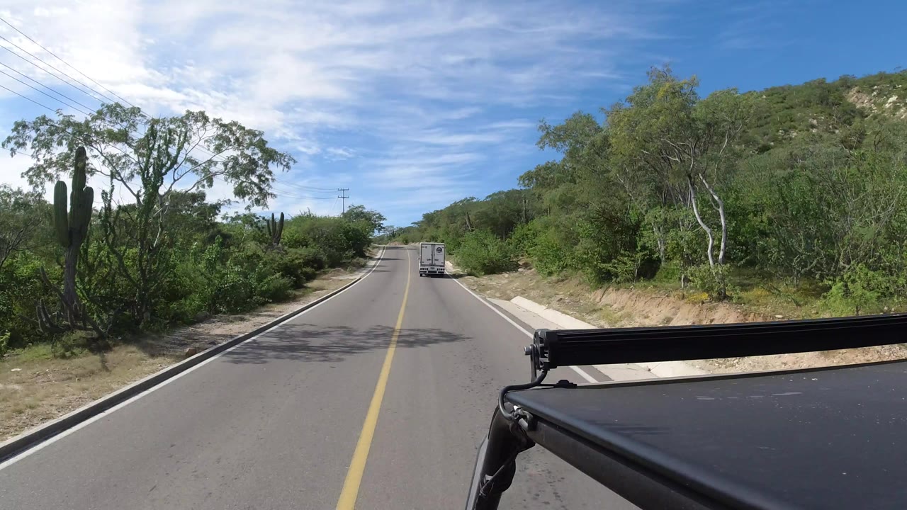 Start of a Los Cabos, Mexico road view with a white box truck ahead, black vehicle bodywork at lower right, roadside trees and cacti, utility lines, and no visible people.
