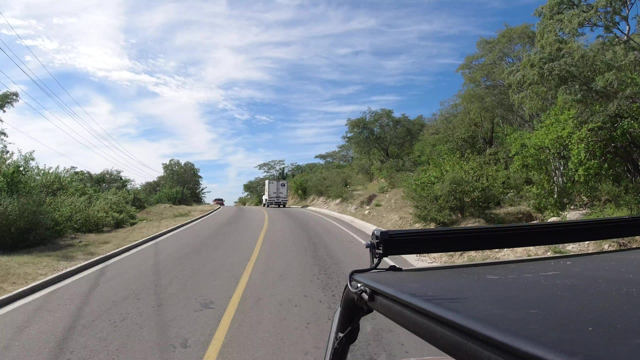 A Los Cabos rural road stretches uphill under bright clouds, with a white truck ahead, green roadside brush, utility wires, and no visible people.