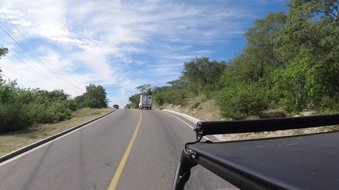The road continues through Los Cabos countryside with a white truck in the lane ahead, a rocky shoulder at right, utility lines above, and no visible people.