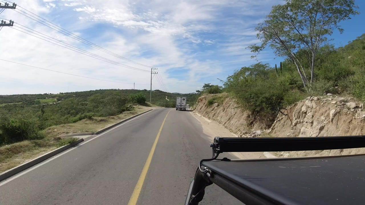 Opening Los Cabos, Mexico road view from a car, with a truck ahead, utility wires above, rocky slope at right, green hills at left, and no visible people.