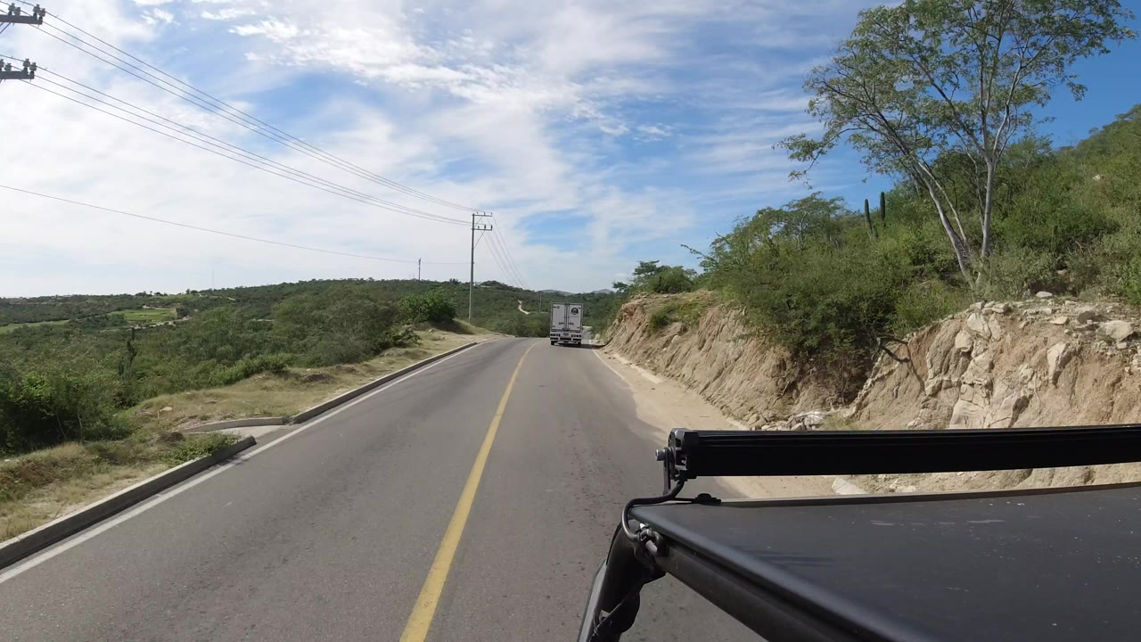 Los Cabos road continues downhill with the truck farther ahead, black car rack in the lower right, power lines over the lane, and no visible people.
