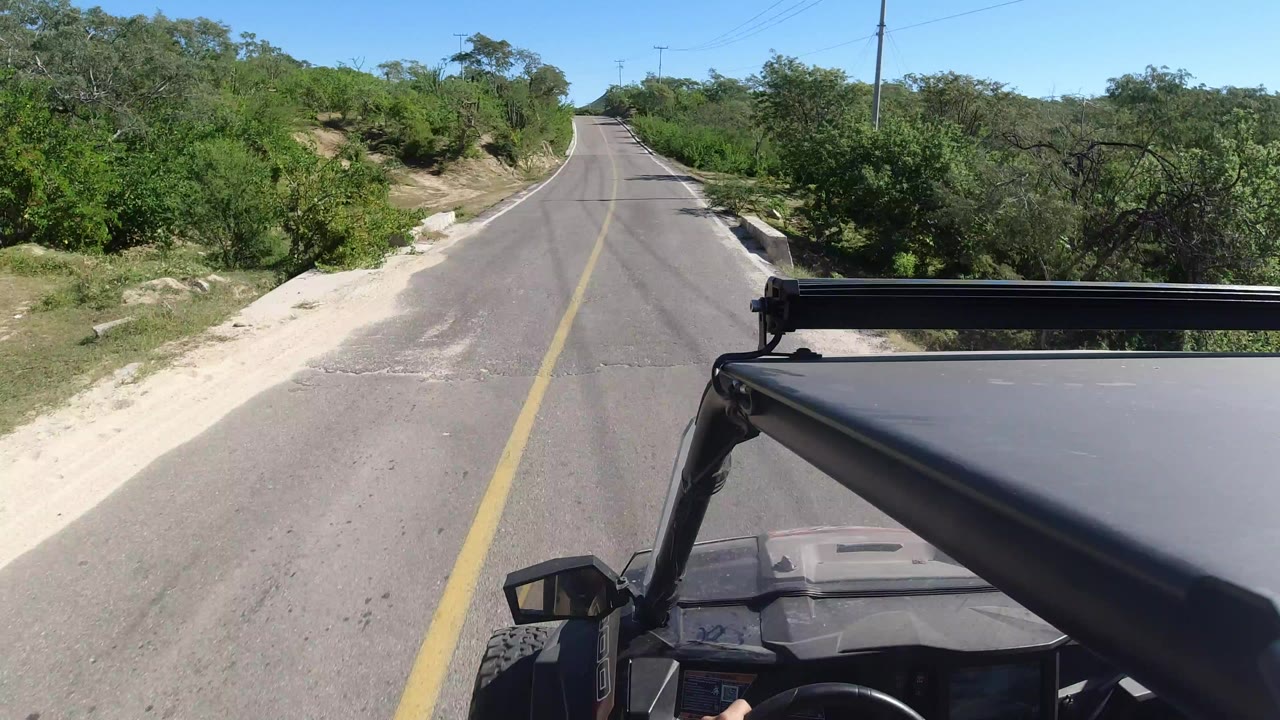 Front-vehicle view of a two-lane road in Los Cabos, Mexico, with a yellow centerline, sandy shoulder, dense green roadside brush, blue sky, and no visible people.