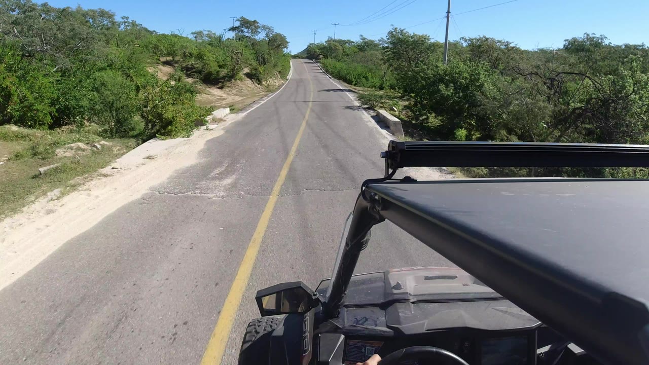 A similar Los Cabos road view continues, with the vehicle corner at lower right, a narrow paved lane ahead, thick green brush on both sides, and no visible people.