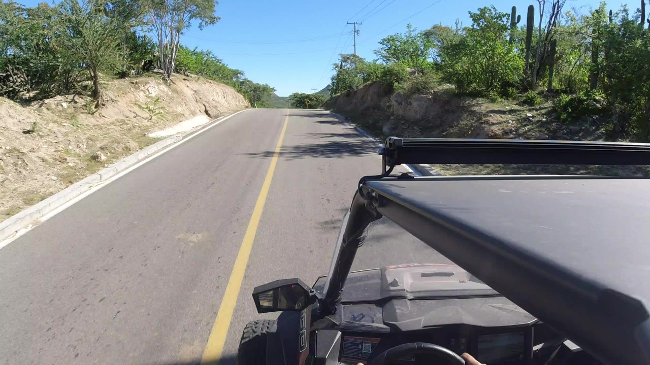 Opening view from a moving vehicle on a paved road in Los Cabos, Mexico, with a yellow center line, sandy roadside banks, dense greenery, and no visible people.