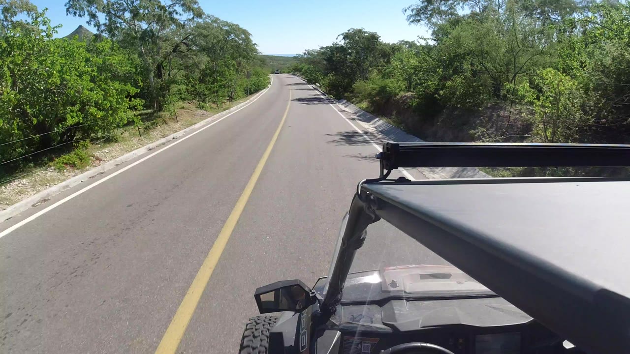 High-mounted jeep view down a two-lane road in Los Cabos, Mexico, with green roadside trees, a yellow center line, and no visible people.