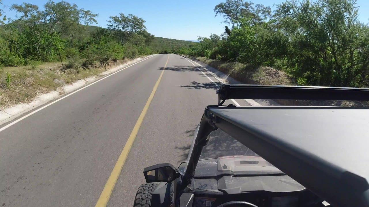 Start of the Los Cabos, Mexico road clip, with a yellow centerline leading forward, vehicle roof hardware in the lower right, green roadside brush, distant hills, and no visible people.