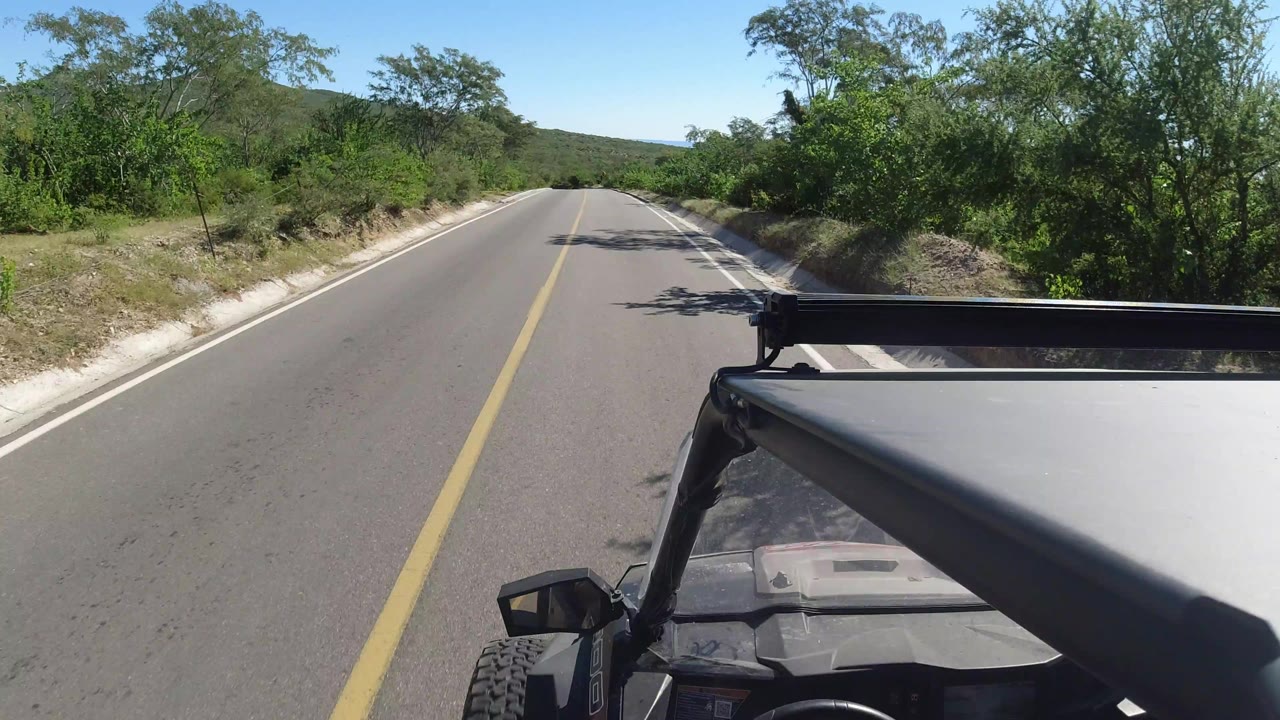 One second into the Los Cabos, Mexico clip, the road remains centered as the safari vehicle foreground fills the lower right, with dense green scrub and low hills under clear light, and no visible people.