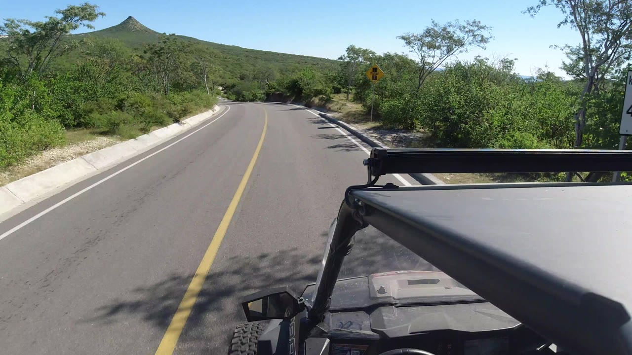 Forward vehicle view on a paved road in Los Cabos, Mexico, with a yellow center line, roadside brush, distant hill peak, and no visible people.