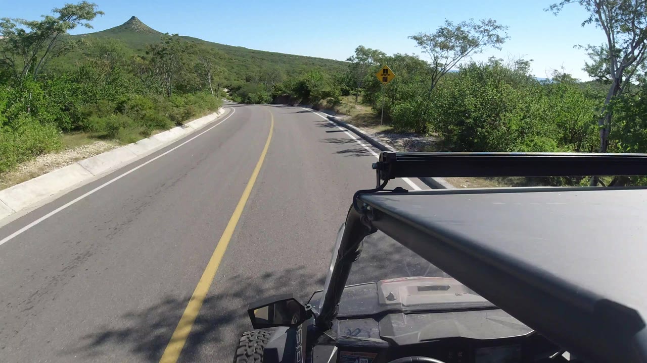 The vehicle continues along the Los Cabos road, with the center line leading toward a shaded bend, dense brush on both sides, and no visible people.