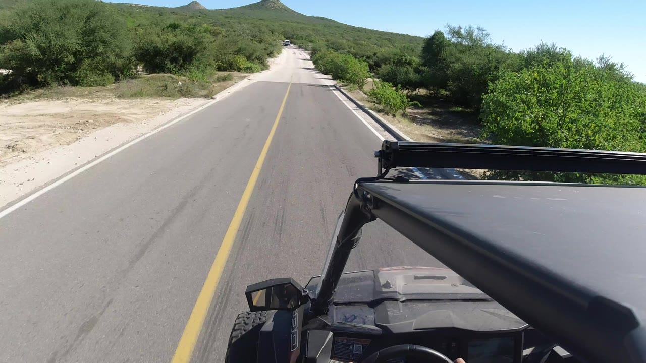 Road ahead through green desert brush with vehicle canopy in foreground.