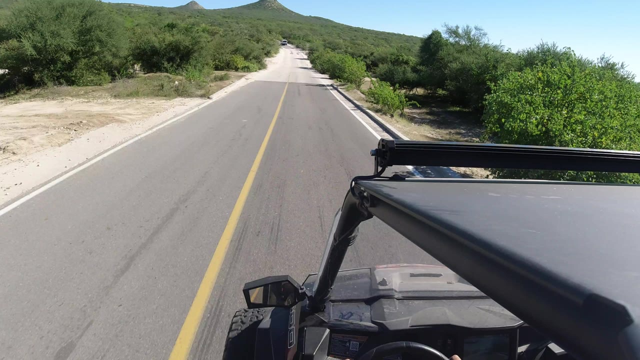 A centered Los Cabos roadway stretches toward low hills, bordered by dense green scrub, with the off-road vehicle canopy and mirror in the foreground and no visible faces.