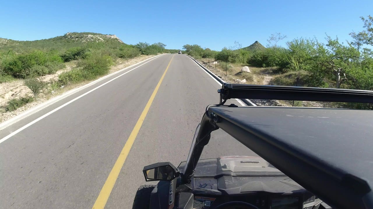 Start of a Los Cabos road drive, showing a paved lane, yellow center line, green brush, low rocky hills, blue sky, and no visible people.