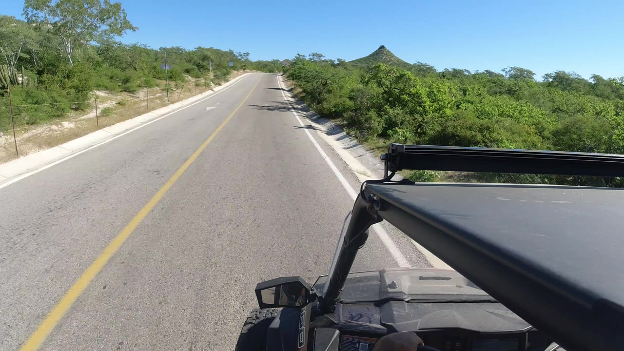 A Los Cabos two-lane road stretches ahead under a clear blue sky, with a vehicle roof and black roll bar in the lower foreground and no visible people.