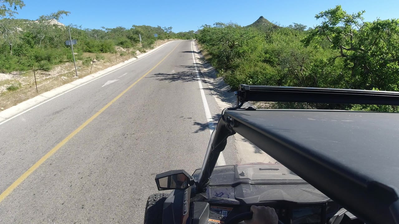 Second frame shows the same Los Cabos roadside view, with the SUV roof and mirror lower in frame, the road stretching ahead, green trees to the right, and no visible people.