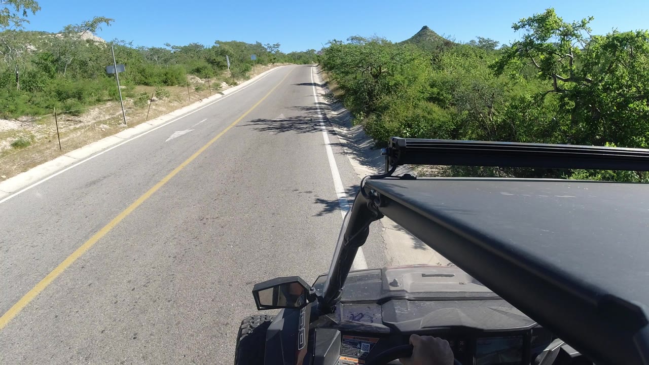 Forward view from an open Jeep on a sunny two-lane road with desert scrub ahead.