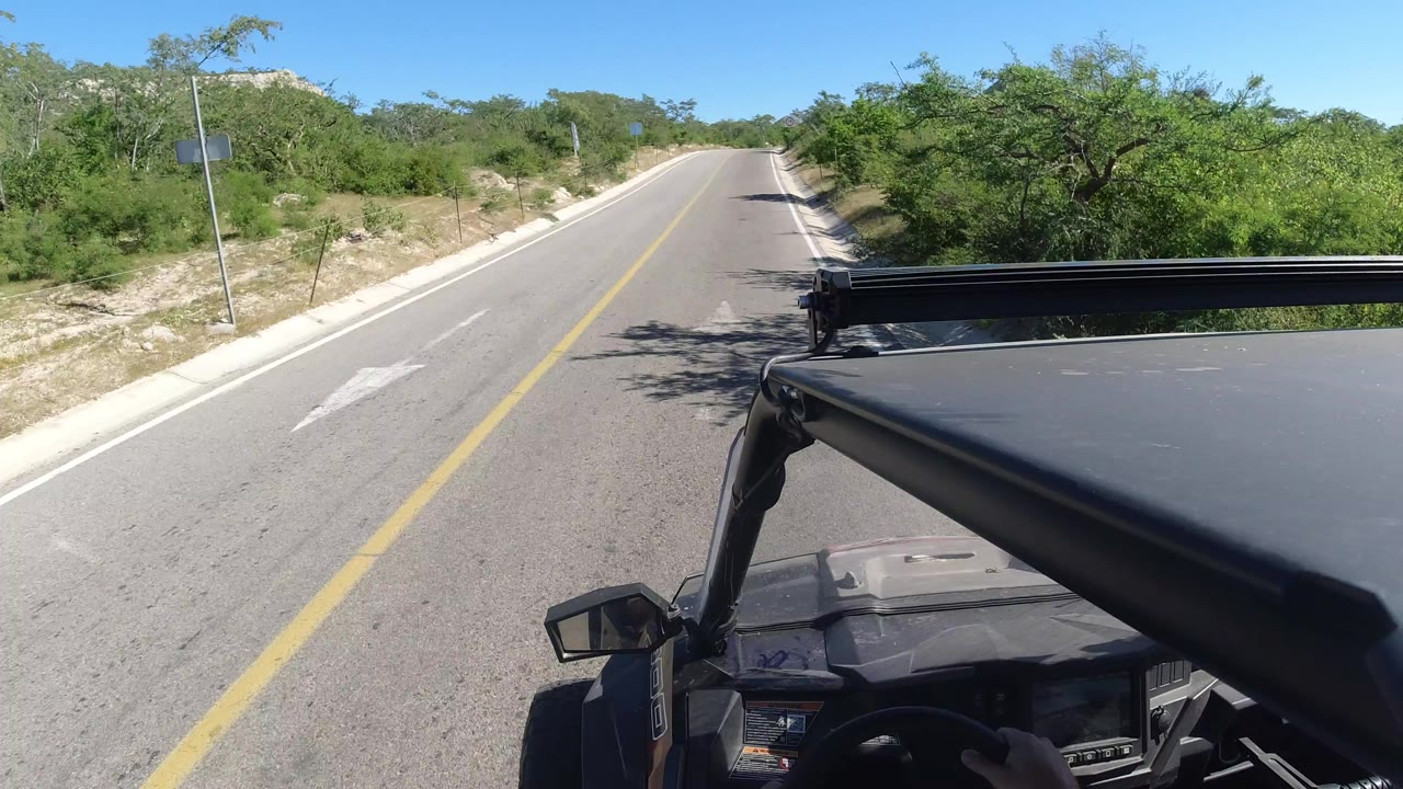 Start of a Los Cabos road POV with a yellow centerline, low roadside vegetation, a sign at left, and vehicle roof hardware in the foreground.