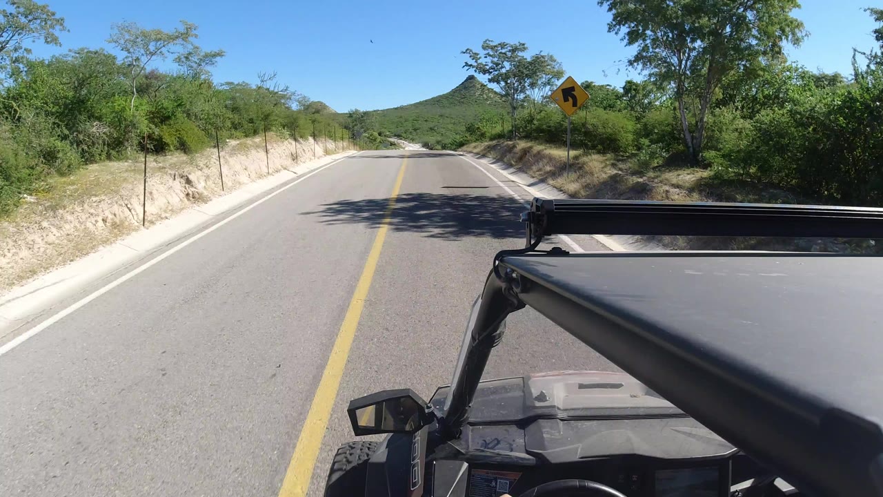 Start of a Los Cabos road POV with a yellow center line, sandy shoulders, green brush, a distant cone-shaped hill, vehicle hardware in the foreground, and no visible people.