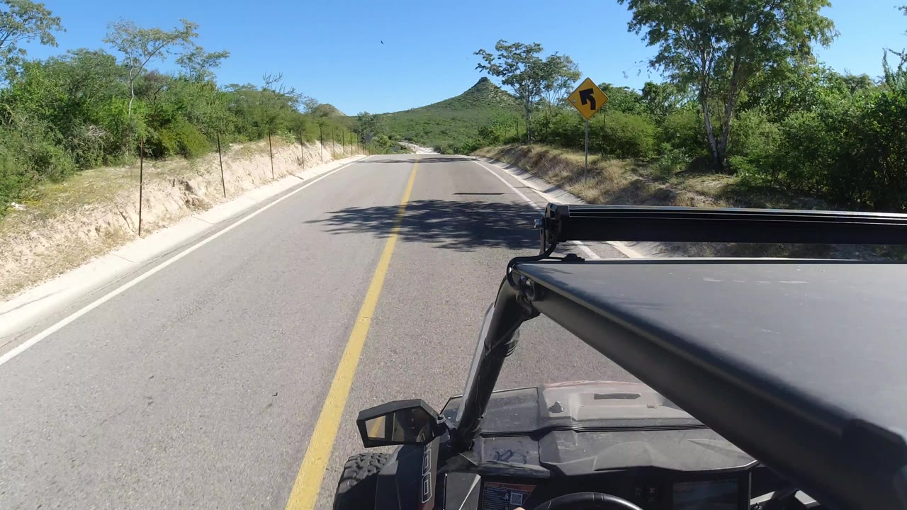 Los Cabos driving view one second in, with the road running between sandy banks, a yellow curve warning sign on the right, dense green roadside vegetation, and no visible people.