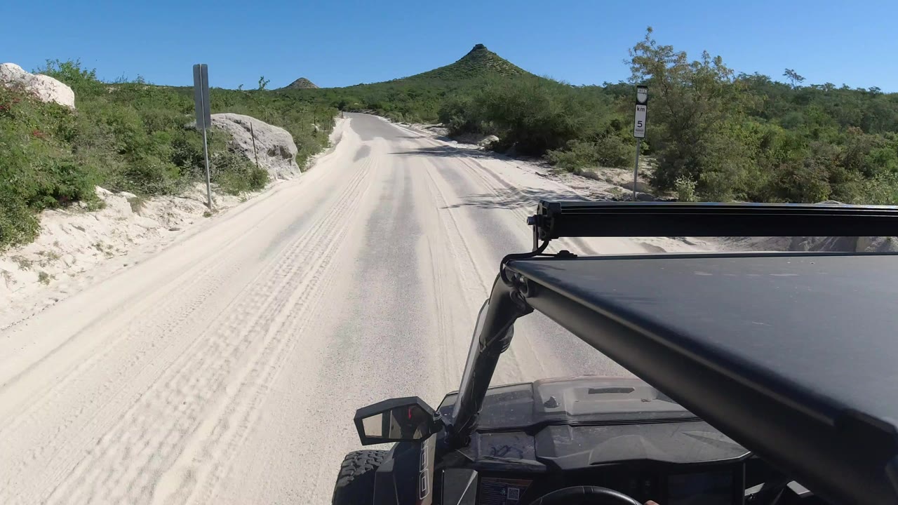 At 0:00 in Los Cabos, the jeep hood and roof bar fill the lower right as a pale sandy road runs toward green scrub and a pointed mountain, with no visible people.