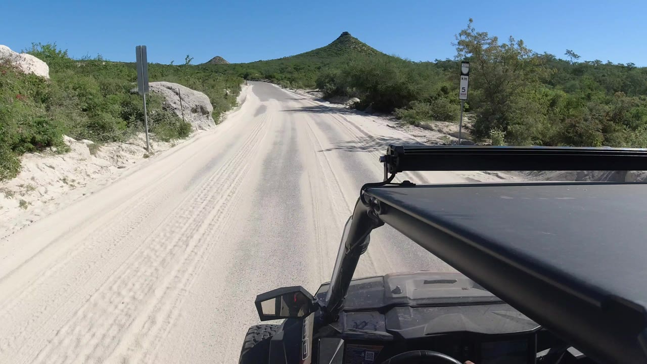 At 0:01 in Los Cabos, the jeep continues along the sandy road between dense green roadside vegetation, pale rocks, and distant hills under clear blue sky, with no visible people.