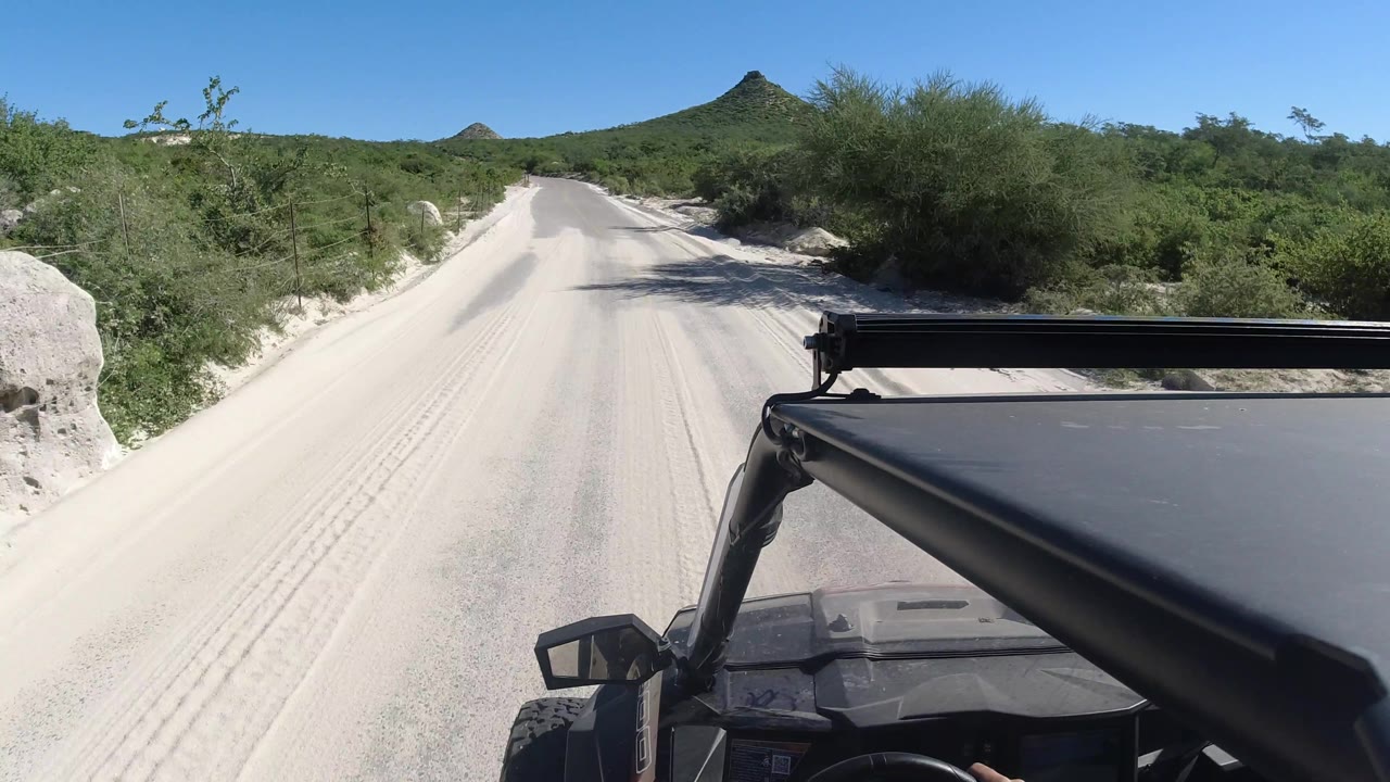 Jeep passes a sandy road curve with shrubs and a mountain on the horizon.
