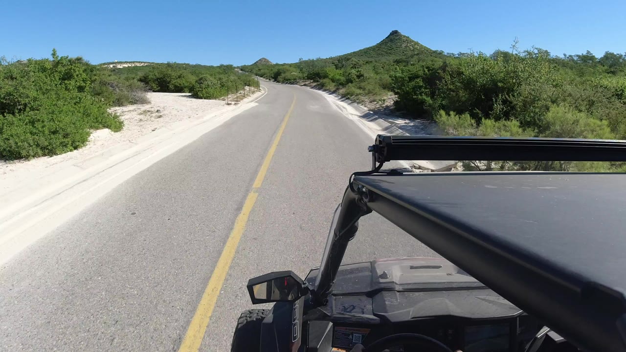 Vehicle-mounted view on a Los Cabos road with a yellow center line, pale sandy shoulders, green brush, a pointed hill ahead, and no visible people.