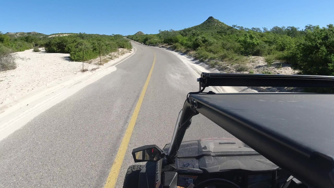 Road curves slightly past sandy shoulder and roadside brush.