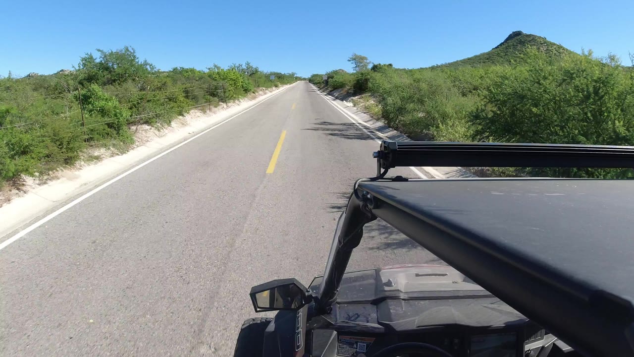 Straight paved road through green roadside brush with vehicle roof rack in foreground.