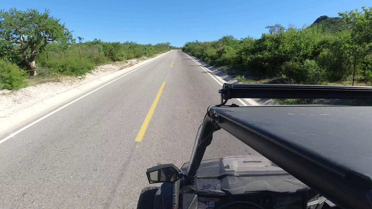 Opening frame of a straight Los Cabos, Mexico road from a vehicle-mounted view, with the hood and side mirror at lower right, green brush along both shoulders, distant low hill, bright blue sky, and no visible people.